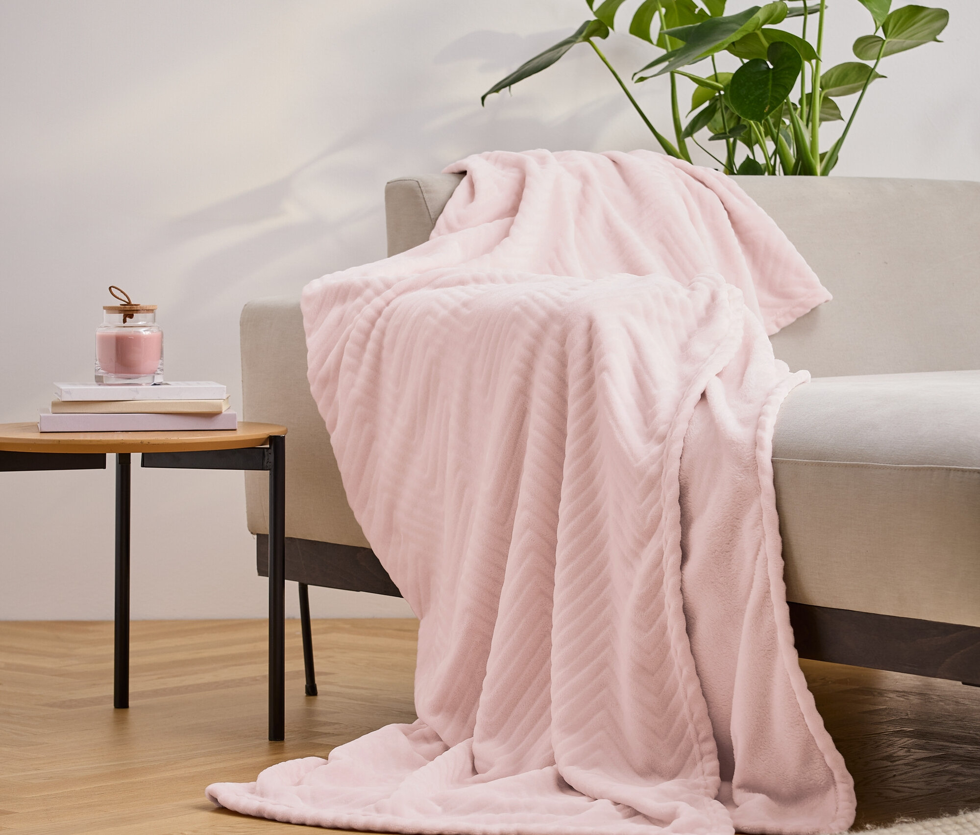 Różowy teksturowany koc narzucony na beżową sofę, obok stolik z książkami i świecą, a w tle roślina.