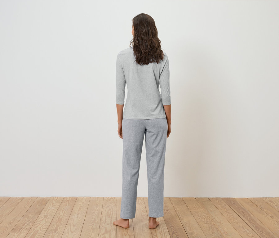 Widok z tyłu kobiety z brązowymi, falowanymi włosami w szarej koszulce z rękawami trzy czwarte i szarych spodniach.