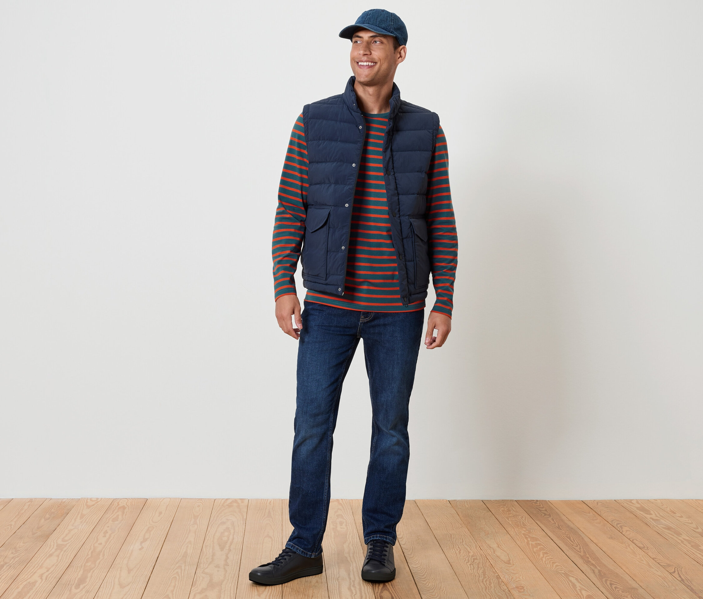 Uśmiechnięty mężczyzna stoi. Ma na sobie niebieską kamizelkę, koszulkę w paski, dżinsy i niebieską czapkę.