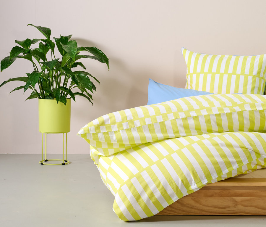 Zielona roślina w okrągłej osłonie na doniczkę stoi obok łóżka z pościelą w żółte paski.