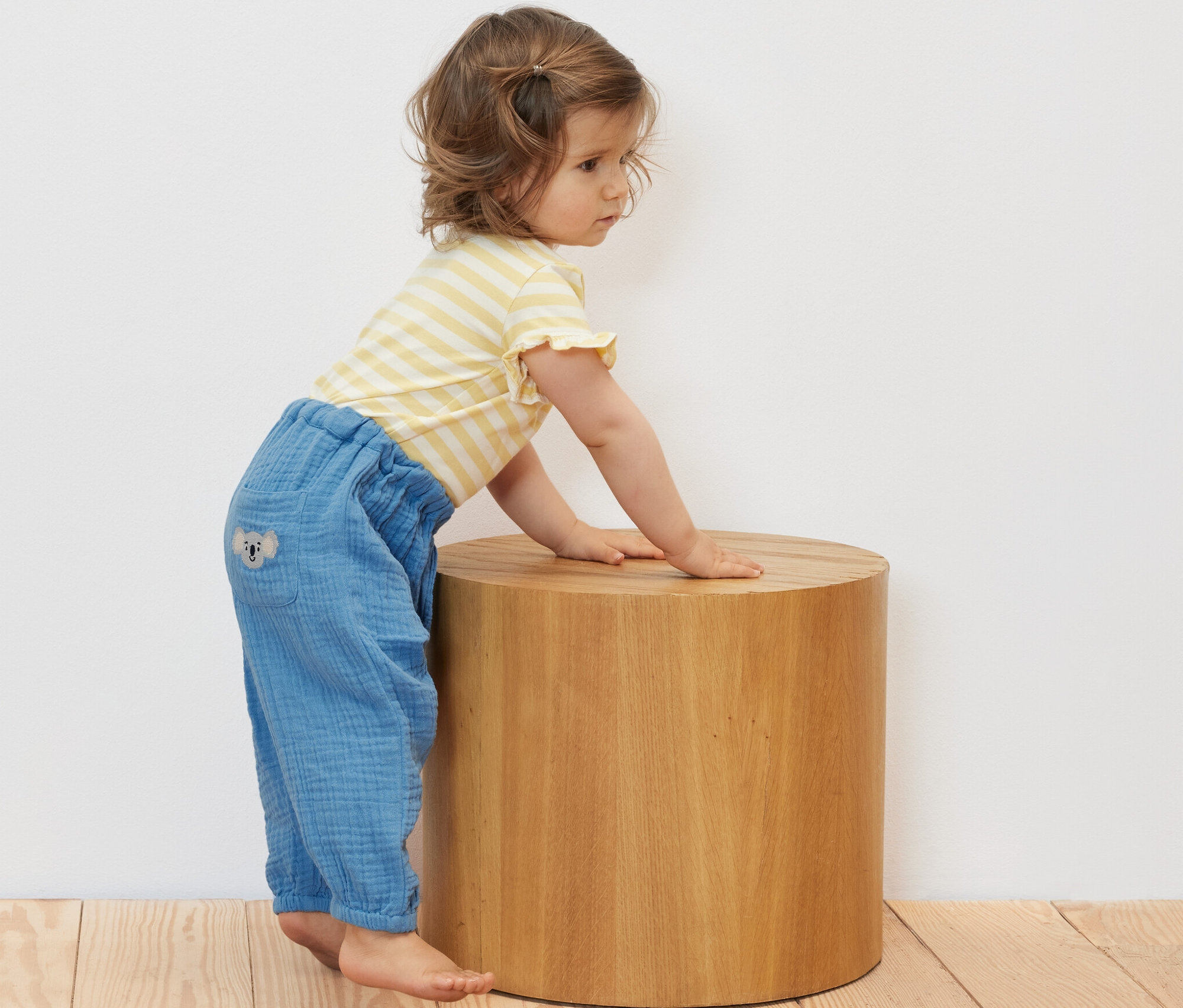 Małe dziecko opiera się o drewniany taboret w muślinowych spodniach na gumce.