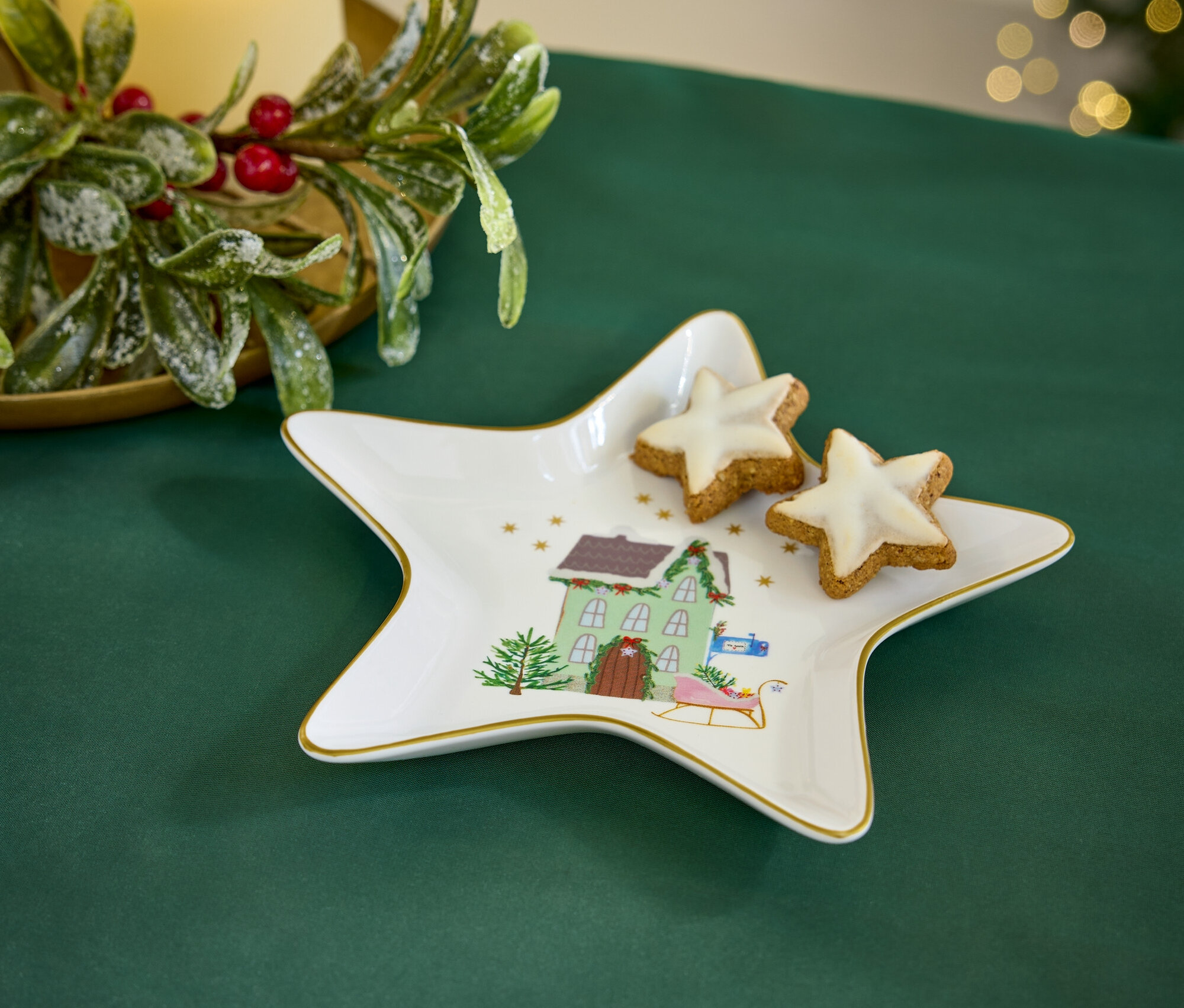 Na zielonym obrusie leży talerz w kształcie gwiazdy z motywem świątecznym i dwa ciasteczka w kształcie gwiazdy. W tle widać świecę i dekoracje świąteczne.