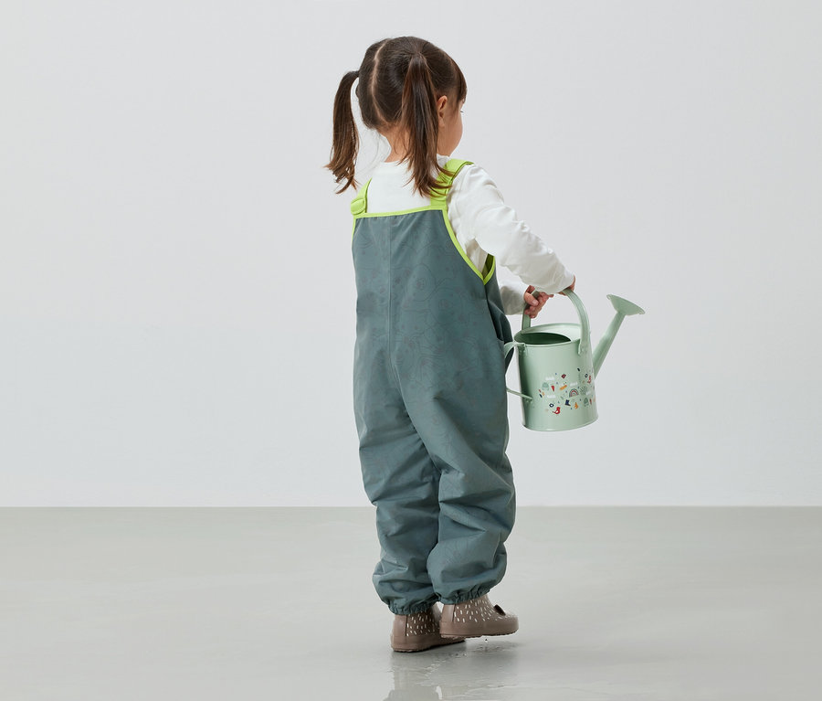 Dziewczynka w szarych spodniach trzyma w ręku zieloną konewkę.