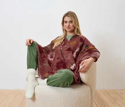 Blondwłosa kobieta siedzi w fotelu, ma na sobie kwieciste kimono, zielone spodnie i białe skarpetki.