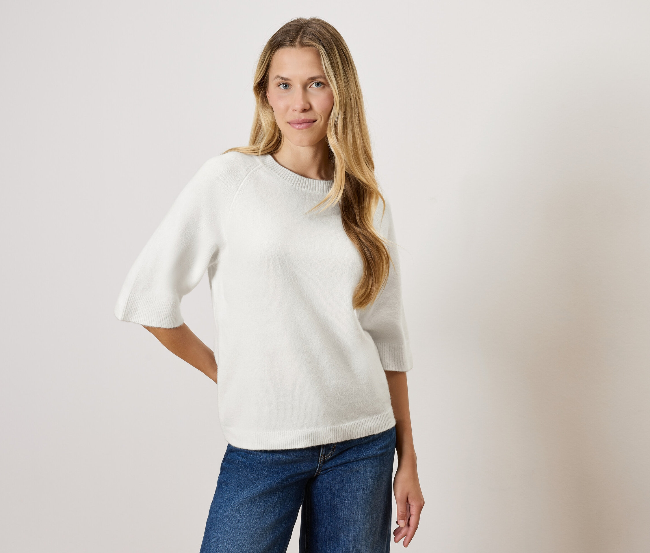 Kobieta z długimi blond włosami pozuje w białym swetrze z rękawami trzy czwarte i niebieskich dżinsach.