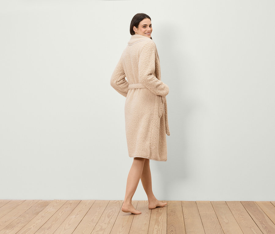 Kobieta spogląda przez ramię, ubrana w szlafrok ze sztucznego futra z paskiem, stojąc na drewnianej podłodze.