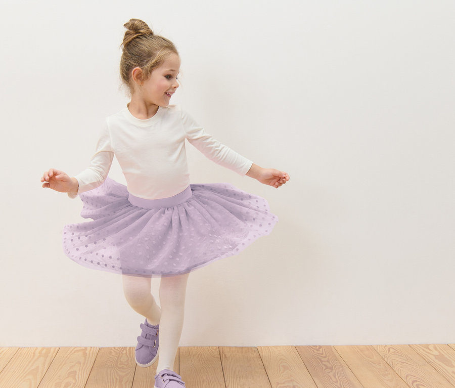 Dziewczynka z kokiem tańczy w liliowej tiulowej spódnicy w białe kropki, białej bluzce z długim rękawem, rajstopach i liliowych butach na tle białej ściany.