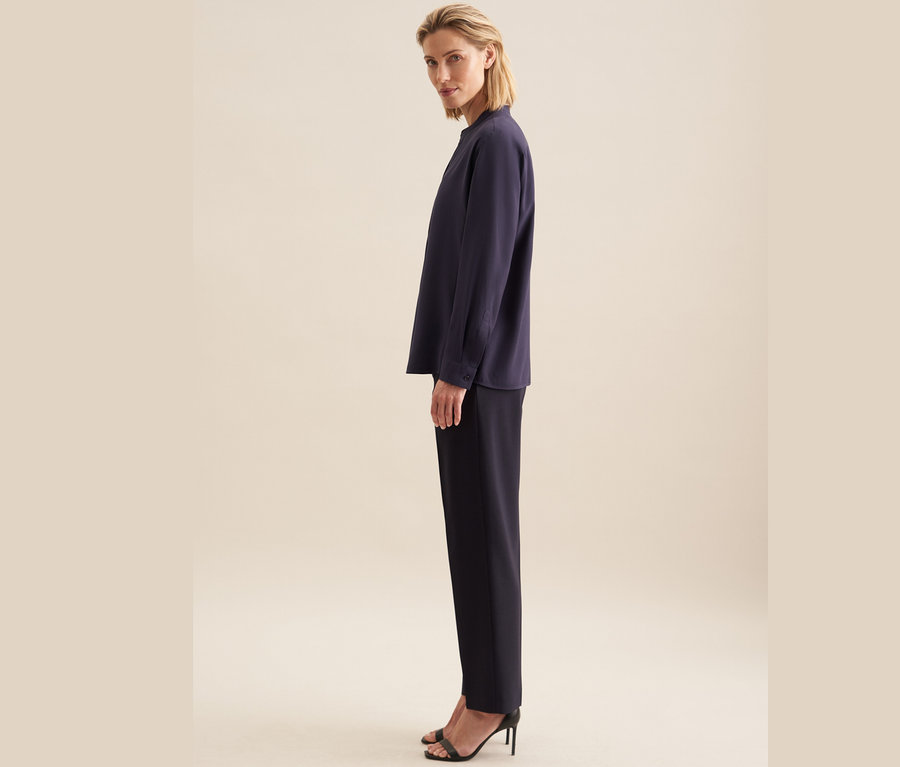 Kobieta stoi bokiem, ubrana w tunikę z wiskozy Seidensticker, czarne spodnie i czarne buty na wysokim obcasie.