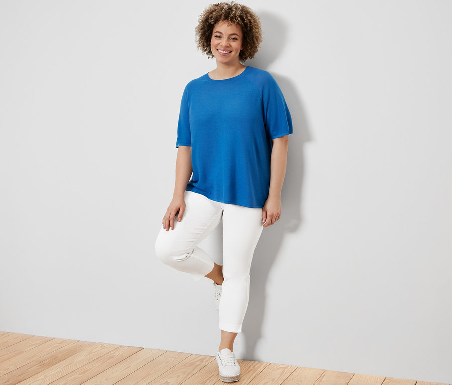 Kobieta pozuje w kobaltowej bluzce z cienkiej dzianiny i białych spodniach stretchowych o długości 7/8.