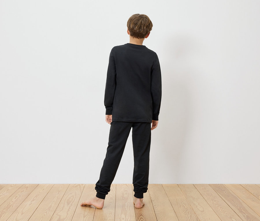 Chłopiec stoi boso na drewnianej podłodze na tle białej ściany, ubrany w czarną piżamę MINECRAFT.