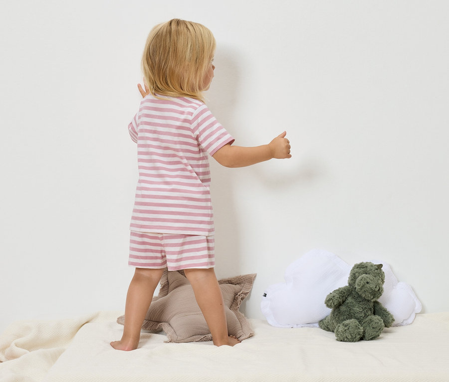 Dziecko z blond włosami w różowo-białej piżamie w paski stoi na beżowym kocu obok zielonego pluszowego dinozaura i białej poduszki w kształcie chmury.