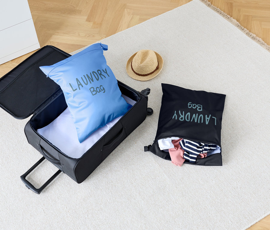 Na dywanie stoi otwarta walizka z niebieskim workiem na pranie w środku, obok czarnego worka na pranie i kapelusza.