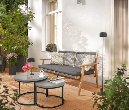 Na tarasie stoją sofa z poduszkami, dwa szare stoliki, lampa podłogowa i rośliny.