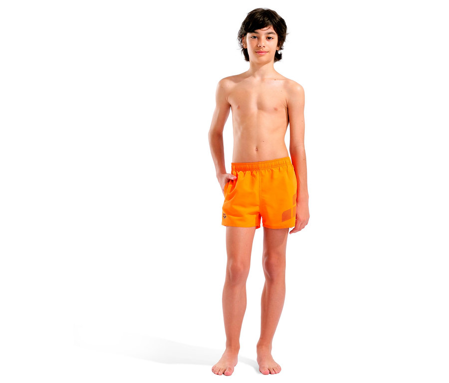 Chłopiec stoi boso na białym tle, ubrany w pomarańczowe kąpielówki.