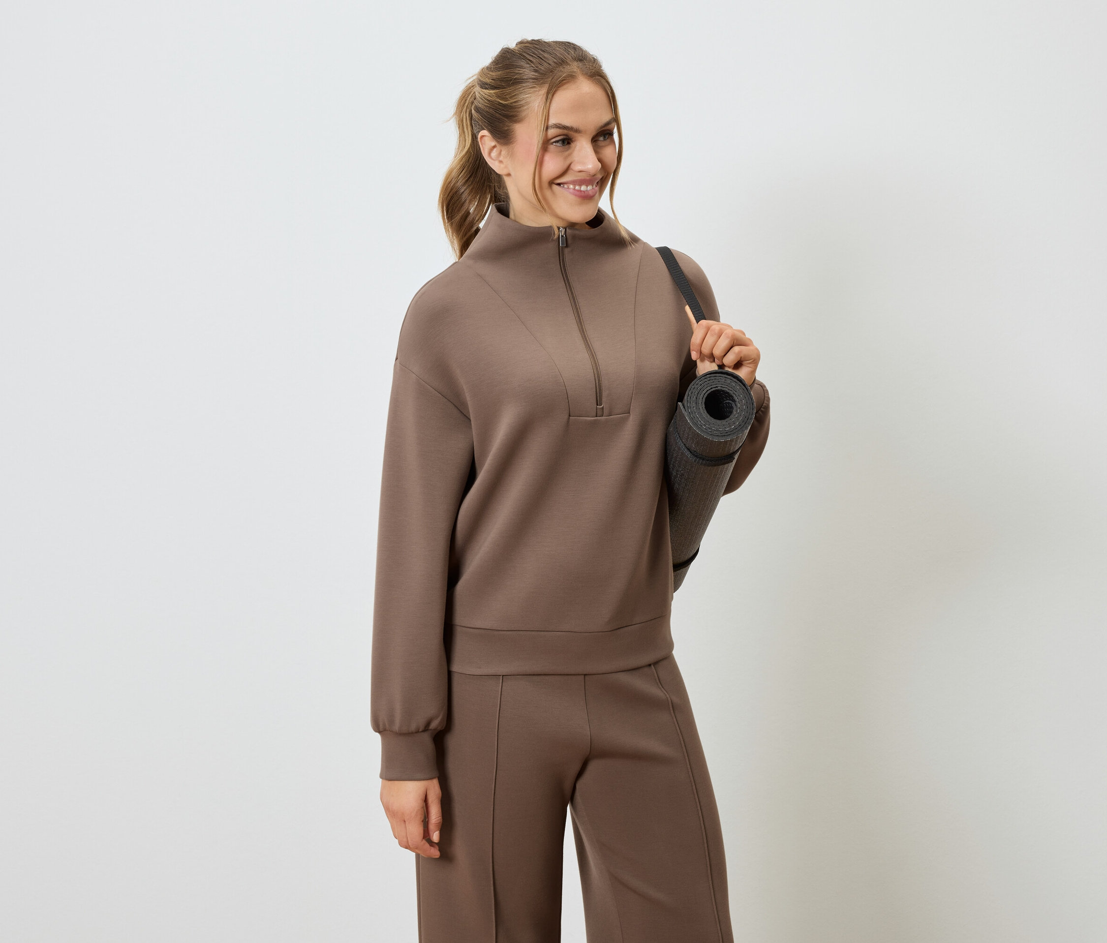 Kobieta w brązowym dresie z matą do jogi na ramieniu.