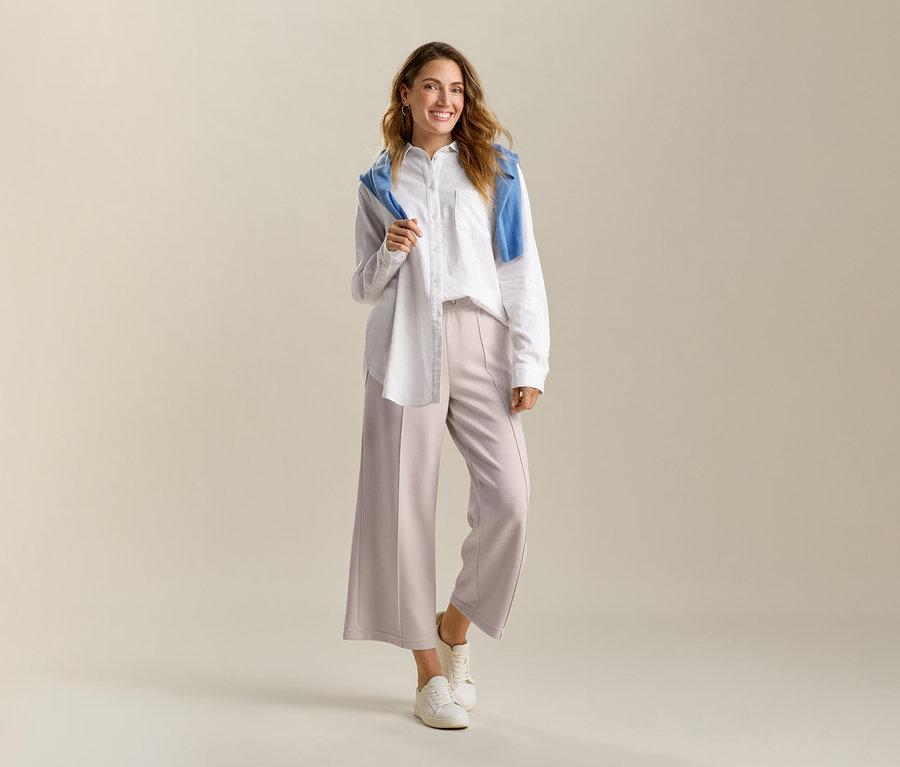 Uśmiechnięta kobieta ma na sobie białą, długą lnianą bluzkę i piaskowe spodnie kuloty z mieszanki modalu. Na ramionach ma niebieski sweter.
