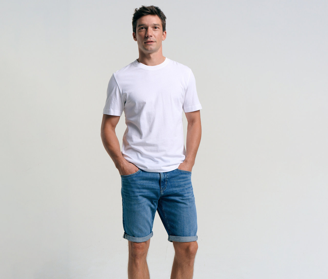 Mężczyzna w białym T-shircie i szortach dżinsowych Tom Tailor TTJOSH REGULAR SLIM, mid stone bright blue denim, z rękami w kieszeniach.