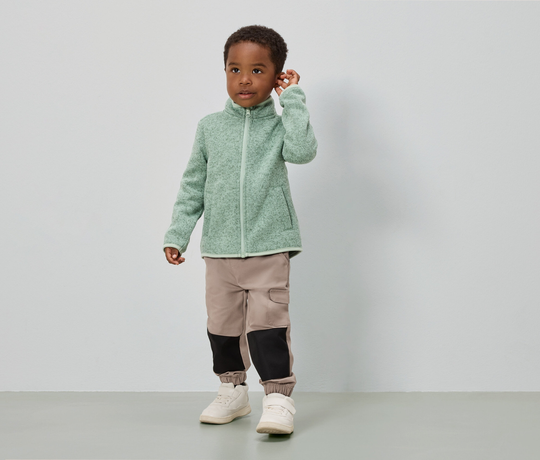 Chłopiec stoi na tle białej ściany, ubrany w jasnozieloną bluzę polarową, beżowe spodnie z czarnymi wstawkami i białe buty.
