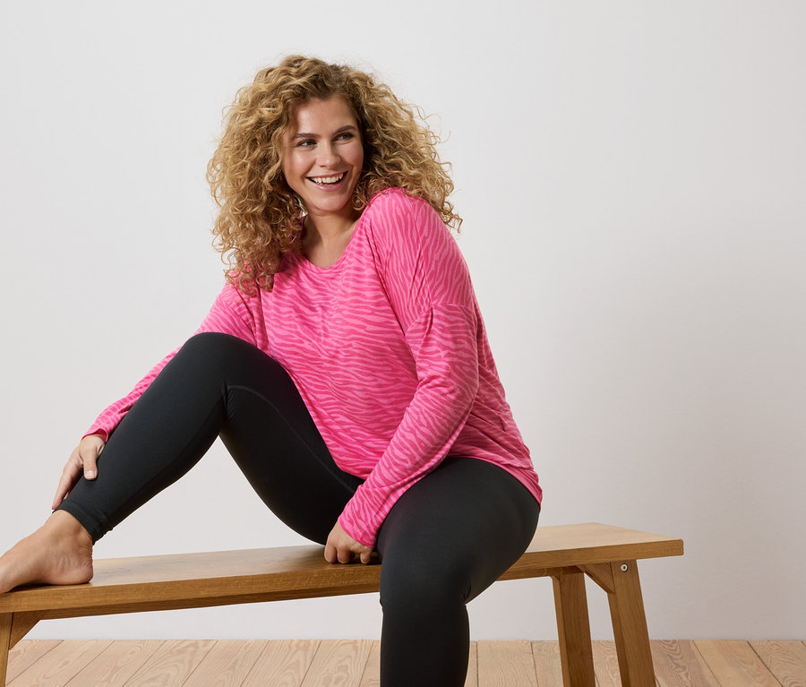 Kobieta z kręconymi włosami siedzi boso na drewnianej ławce, ubrana w różową koszulę z długim rękawem w zebrę i czarne legginsy.