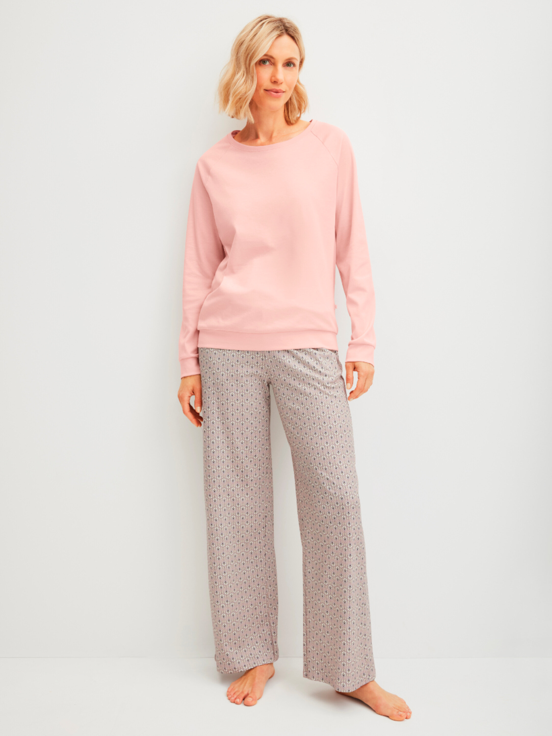 Kobieta stoi boso w różowym swetrze i wzorzystych spodniach na tle białej ściany.