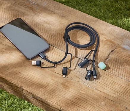 Szary smartfon podłączony do czarnego, plecionego kabla wielokrotnego ładowania leży na drewnianej ławce.