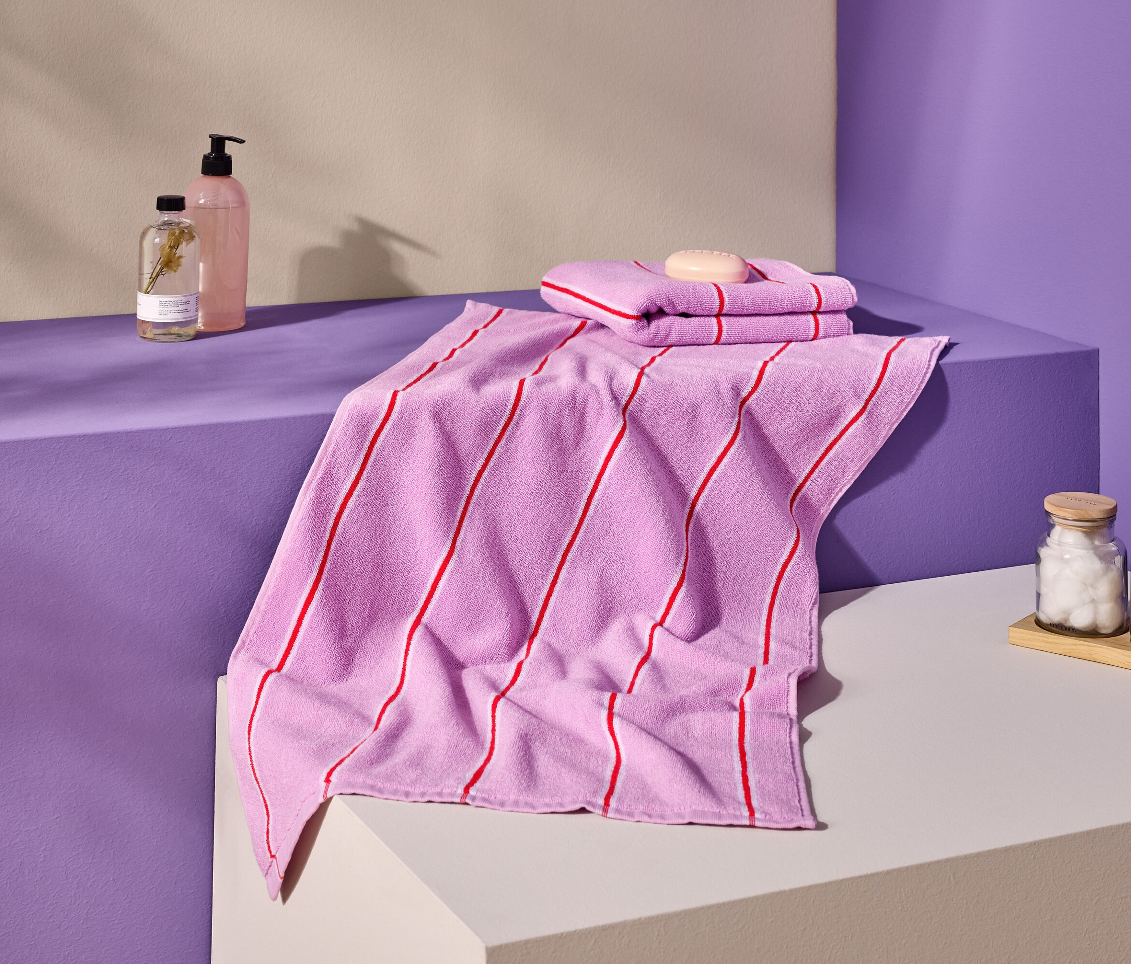 Zdjęcie różowych ręczników w czerwone paski, mydła, butelek i pojemników na fioletowo-kremowej powierzchni.