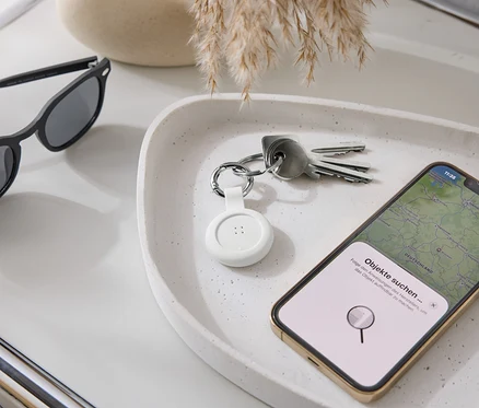 Na tacy leżą okulary przeciwsłoneczne, klucze, biały lokalizator i smartfon z aplikacją map.