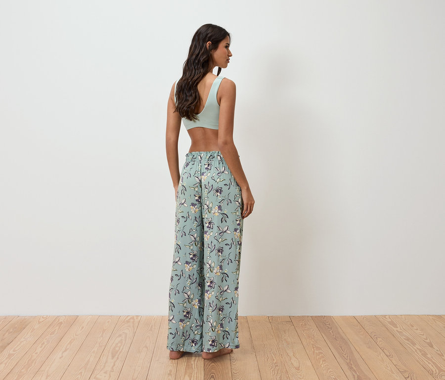 Kobieta stoi tyłem do kamery, ubrana w jasnozielony top i luźne spodnie w kwiaty.