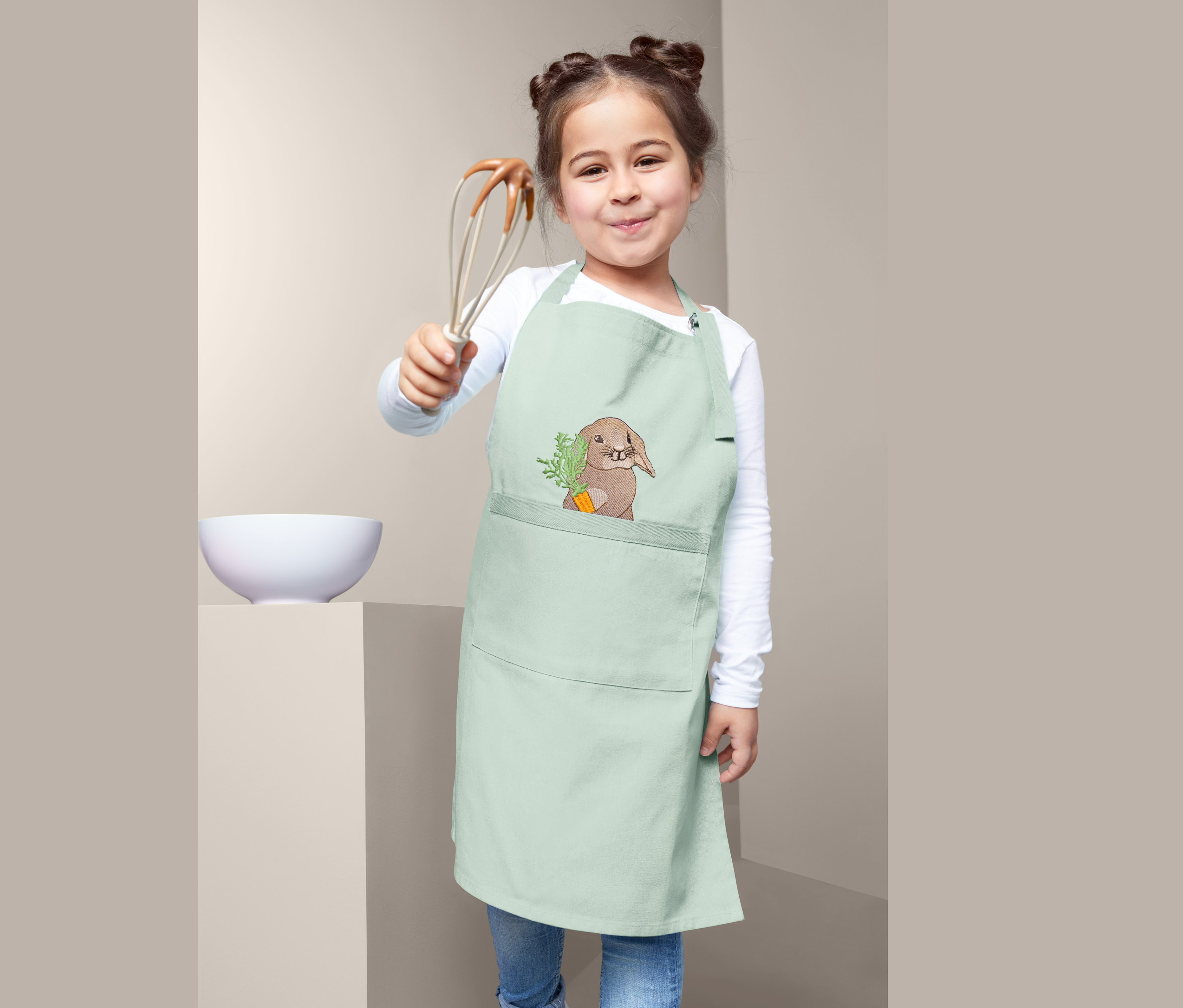 Dziewczynka trzyma trzepaczkę, ma na sobie bawełniany fartuszek dla dzieci, miętowy z motywem zajączka.