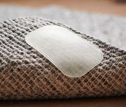 Zbliżenie na róg szarego dywanu z jedną przezroczystą antypoślizgową podkładką pod dywan, 4 sztuki.