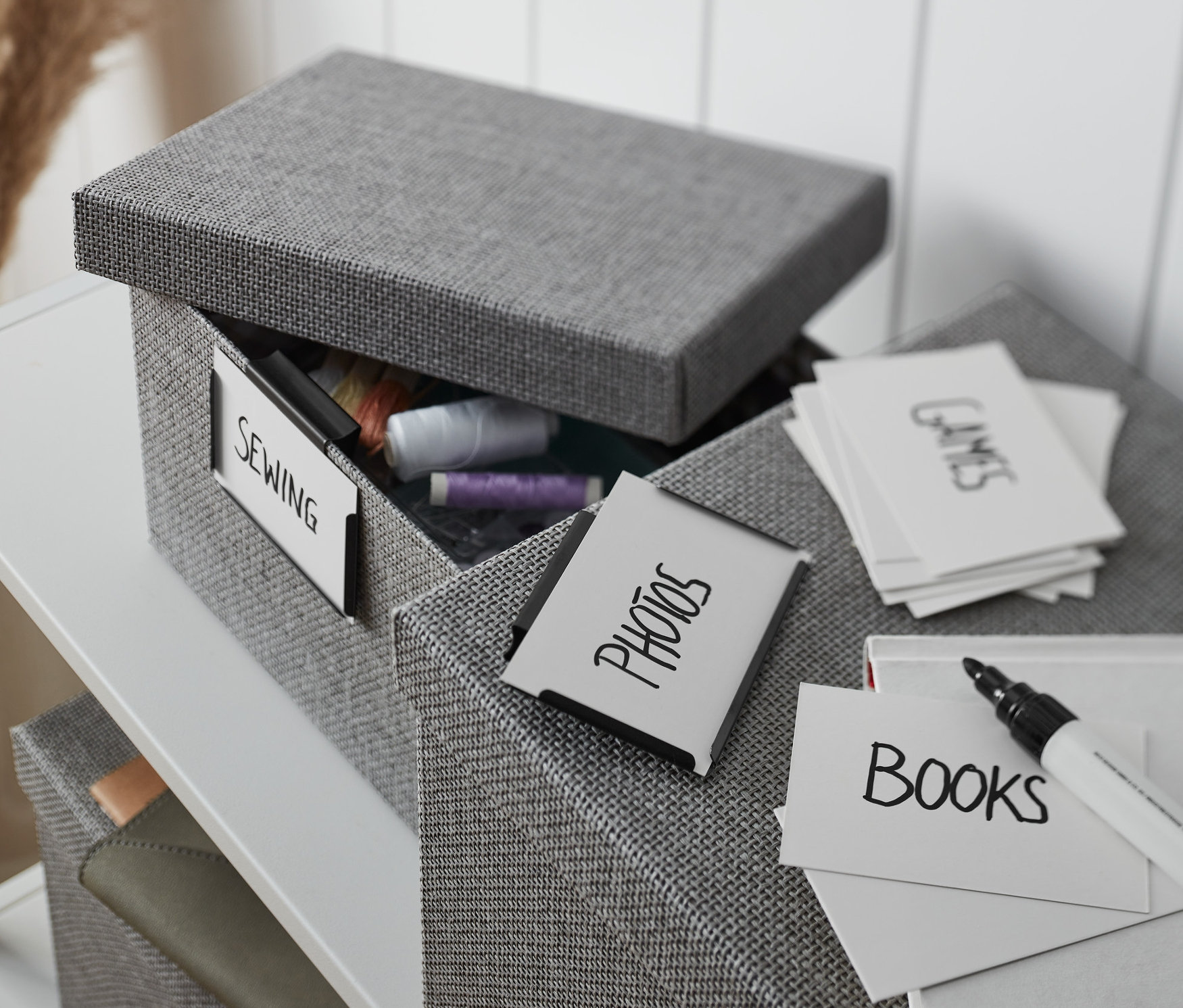 Otwarty szary materiałowy pojemnik z klipsami metalowymi, etykietą „szycie” i zawartością nici, obok pojemnik z kartami z napisami.