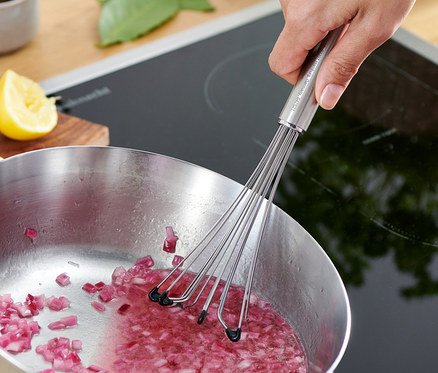 Ręka miesza profesjonalną trzepaczką posiekaną czerwoną cebulę w garnku na kuchence.