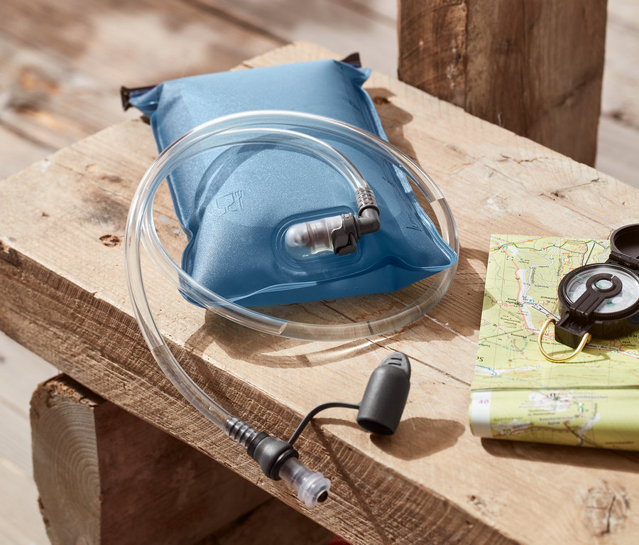 Niebieski bukłak, 2 l, z rurką leży na drewnianym stole obok kompasu i mapy. W tle widać plecak.