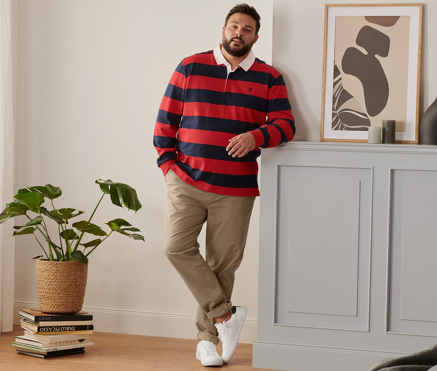 Mężczyzna oparty o ścianę ma na sobie buty typu sneaker, karmelowe spodnie chino i granatowo-czerwoną koszulkę polo z długim rękawem.
