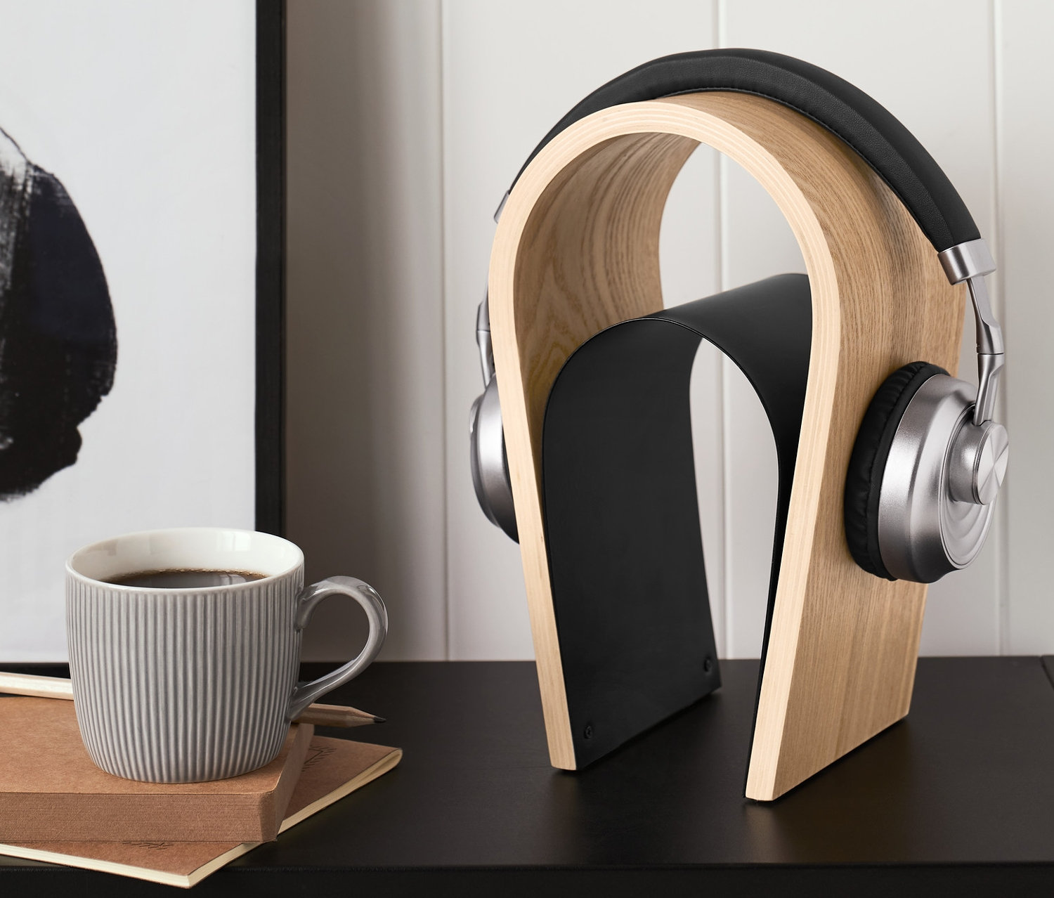 Słuchawki na drewnianym stojaku na słuchawki. Obok stoi szary kubek i leży notatnik.