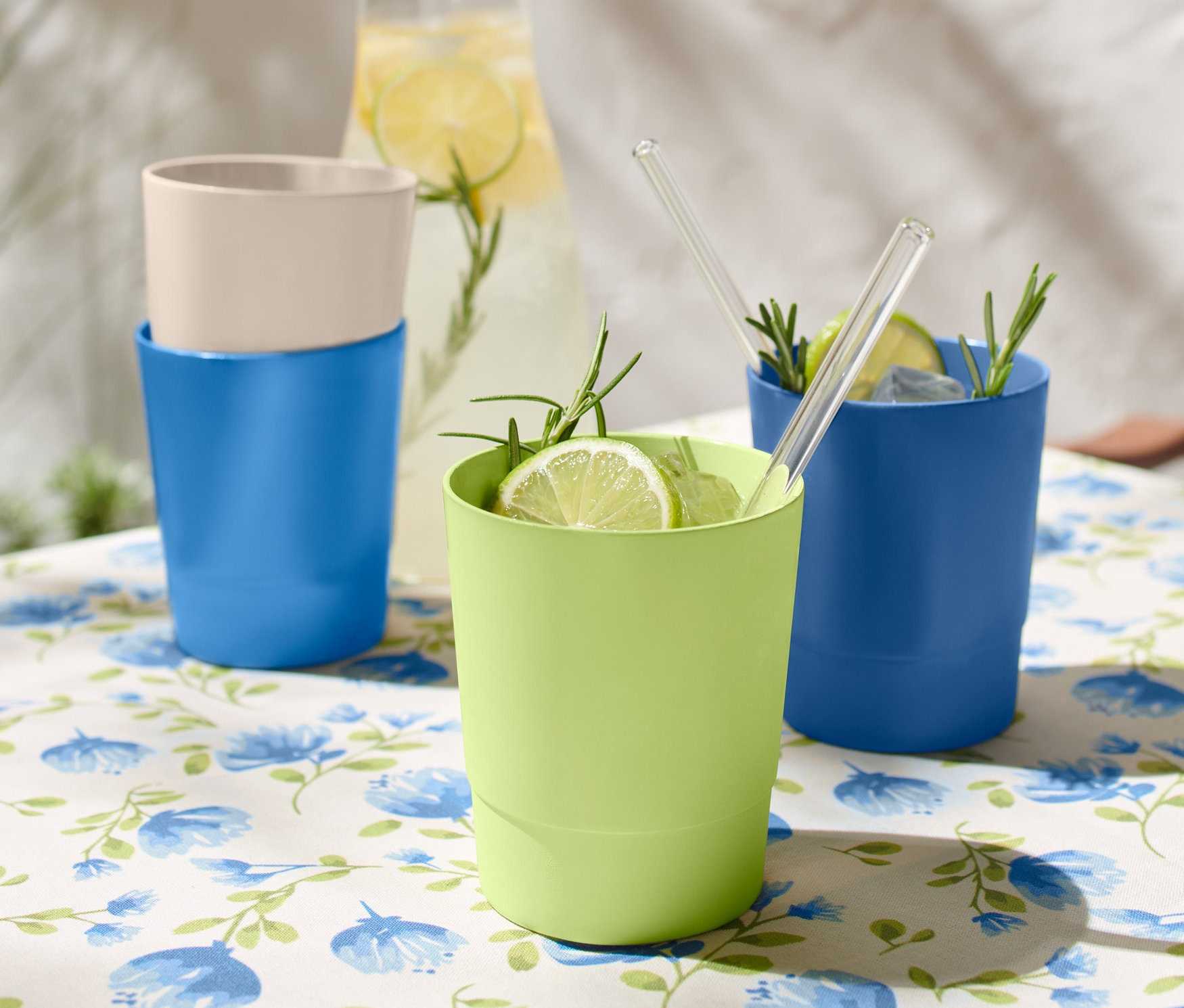 Na stole z kwiatowym obrusem stoją 4 kubki do picia z tworzywa sztucznego, dwa niebieskie, jeden zielony i jeden beżowy.