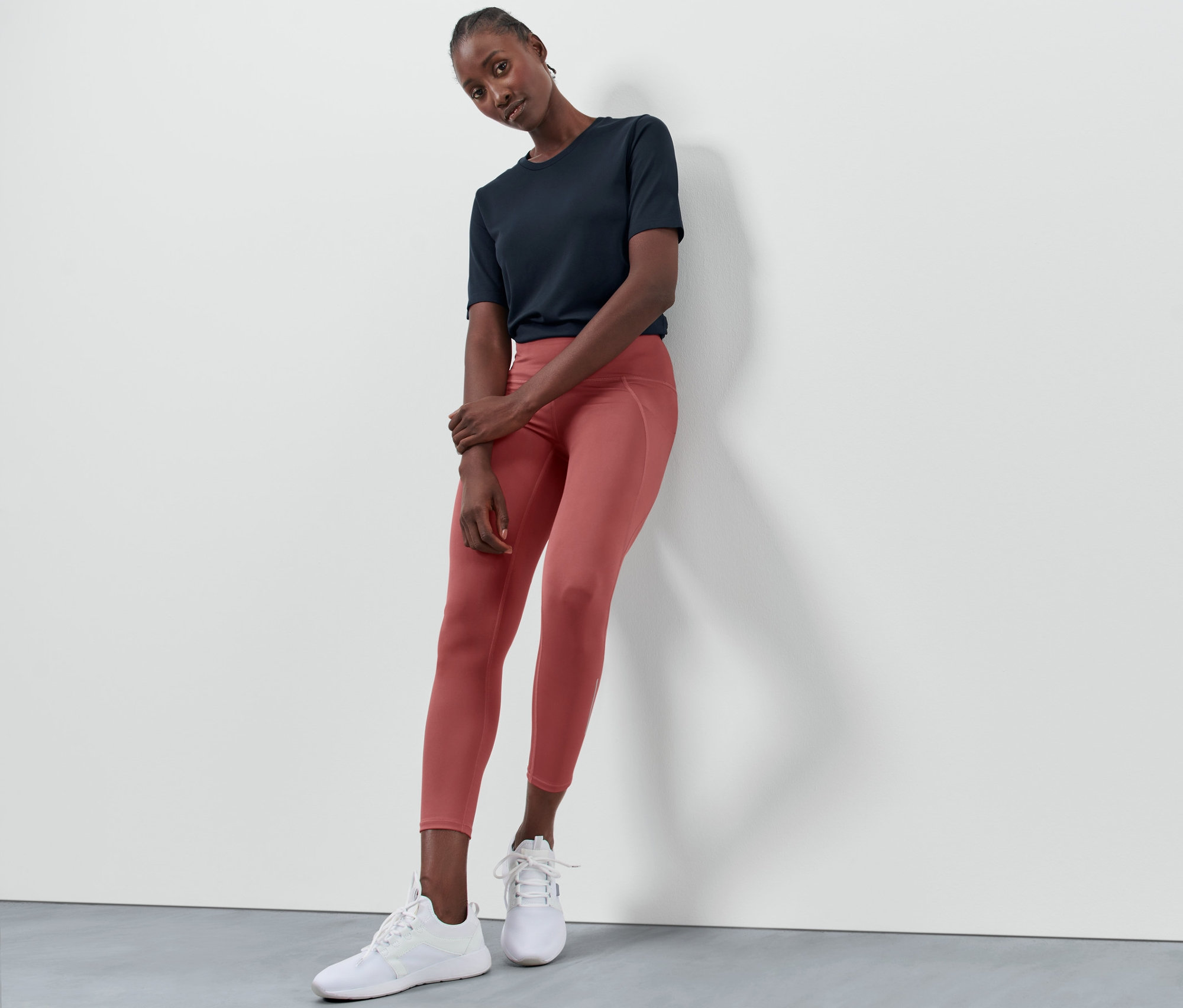 Kobieta oparta o ścianę ma na sobie legginsy sportowe w kolorze terakoty, granatowy T-shirt i białe tenisówki.