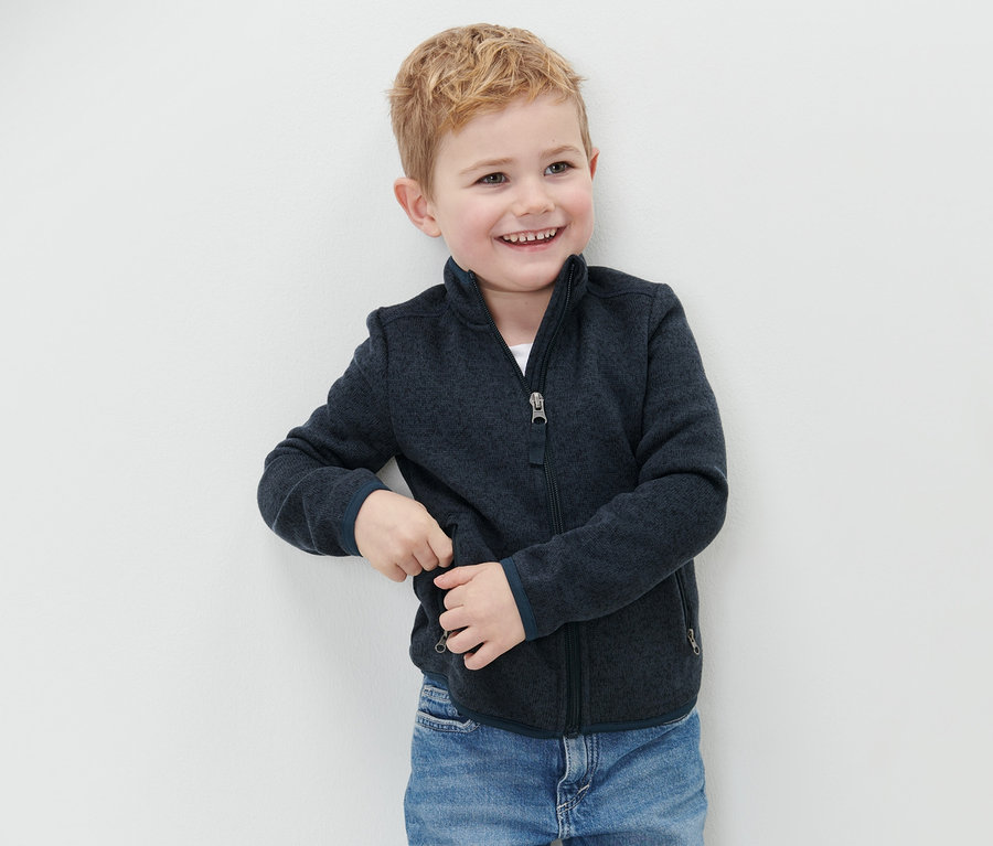 Uśmiechnięty chłopiec stoi przy ścianie w dziecięcej bluzie z dzianiny polarowej i niebieskich dżinsach.