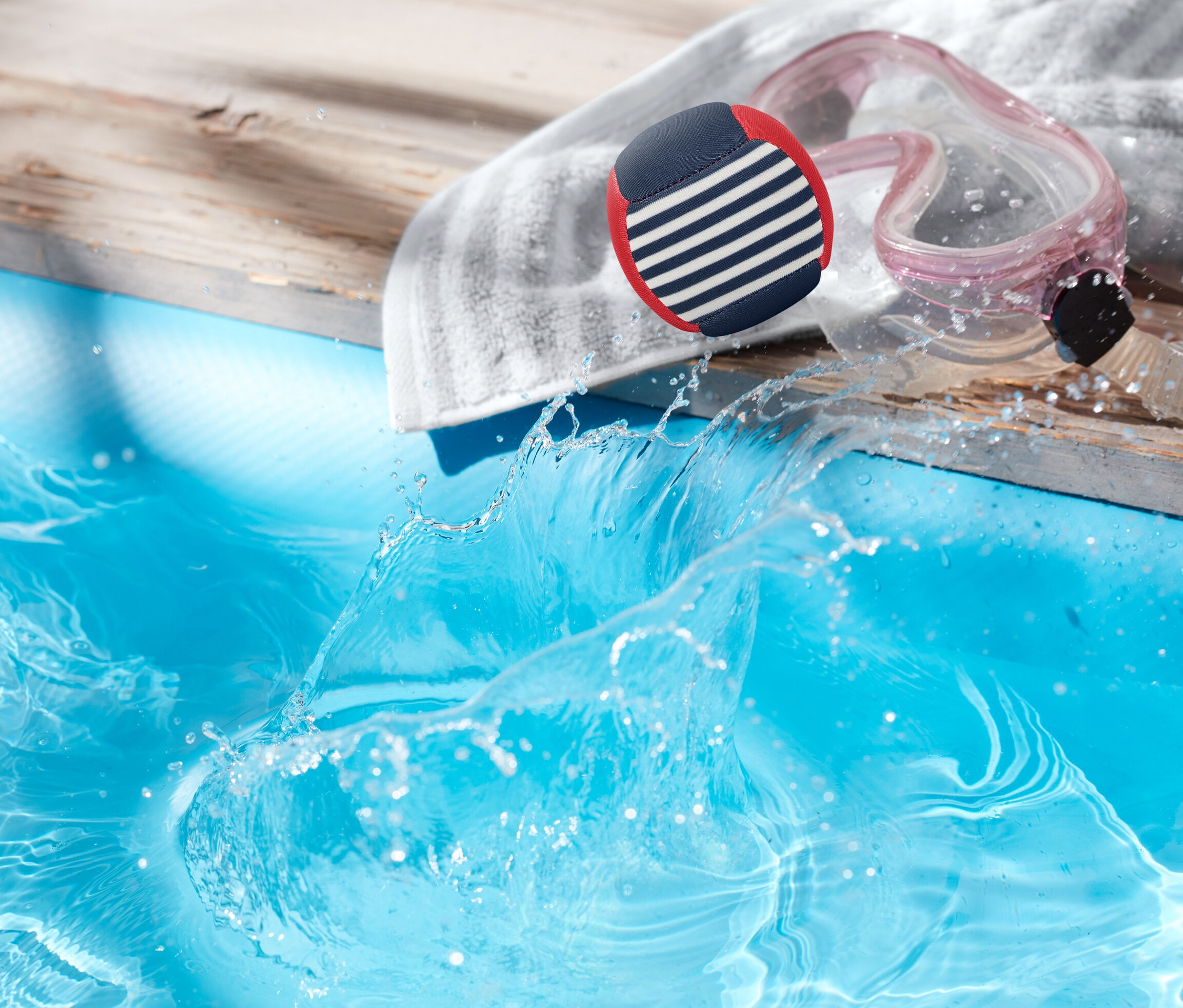 Woda rozpryskuje się w basenie obok drewnianego pokładu, na którym leżą ręcznik, okulary pływackie i niebiesko-biało-czerwona piłka.