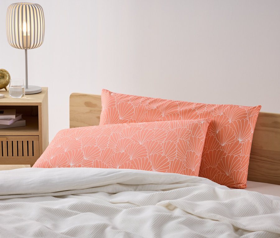 Dwie koralowe poszewki na poduszki z mikrowłókna z białym kwiatowym wzorem leżą na łóżku. Obok łóżka stoi szafka nocna z lampą.