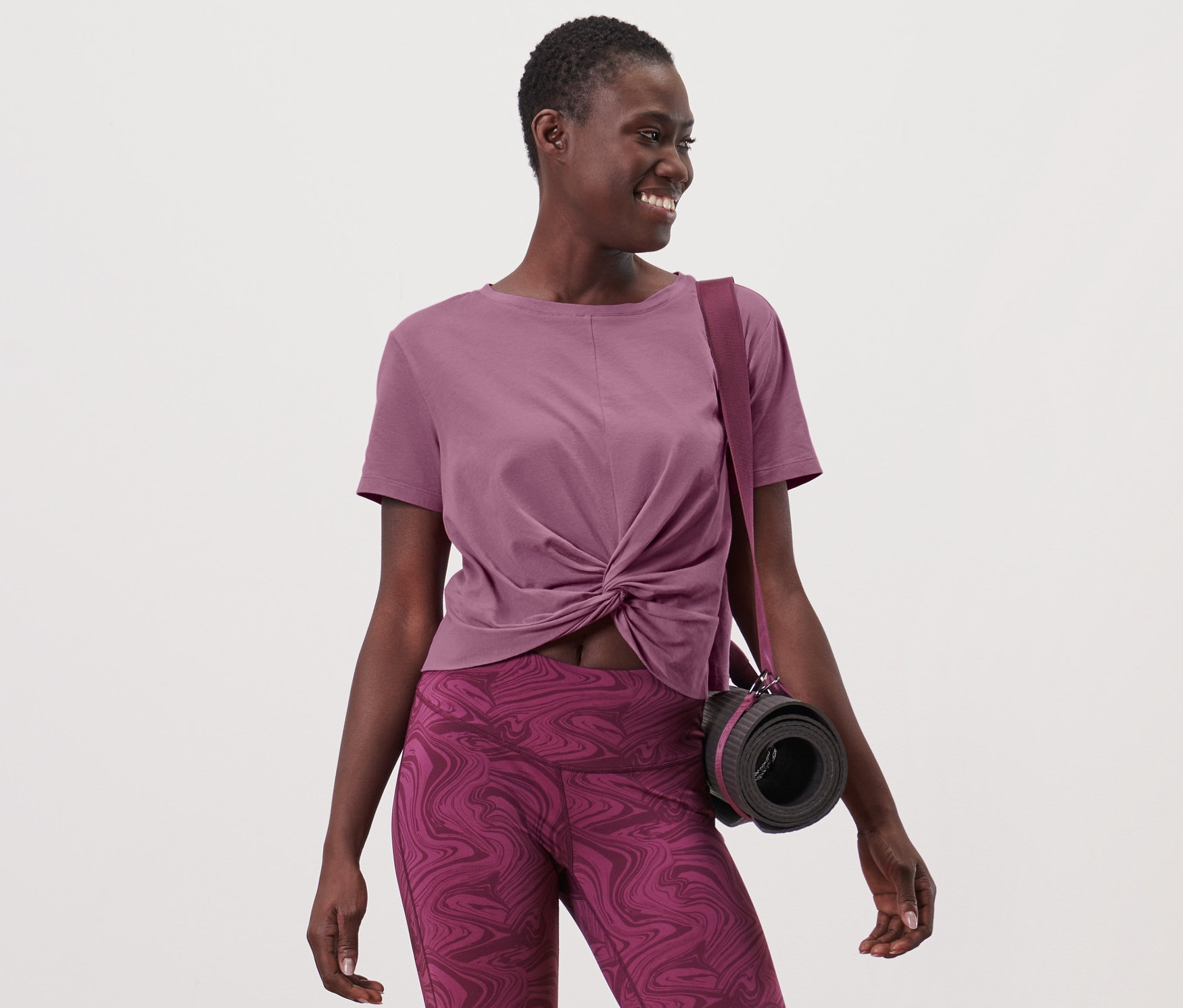 Kobieta ma na sobie koszulkę sportową i legginsy, z torbą i matą do jogi przewieszoną przez ramię.