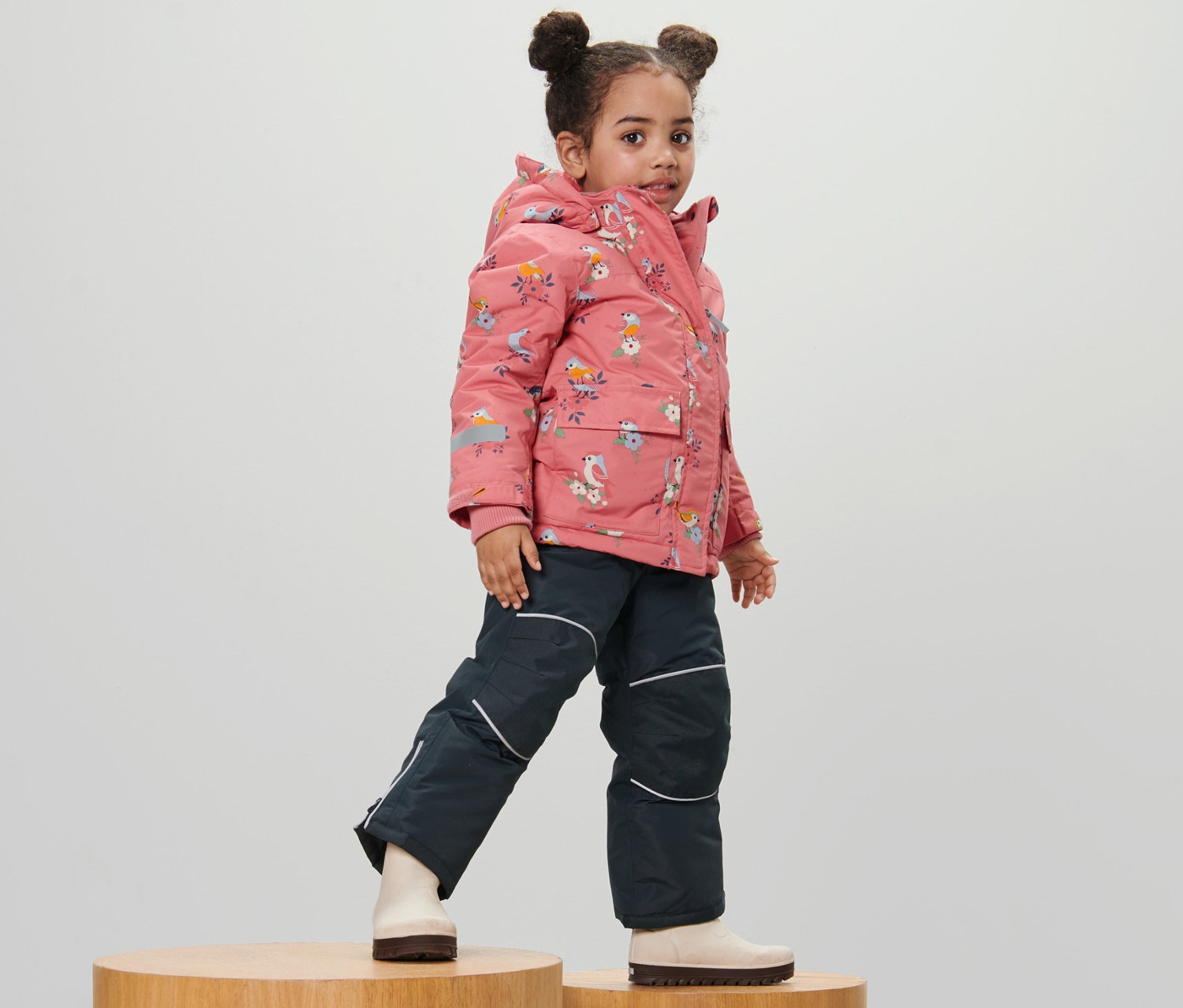 Dziewczynka pozuje na drewnianych postumentach w 2-częściowym kombinezonie zimowym z nadrukiem w ptaki.