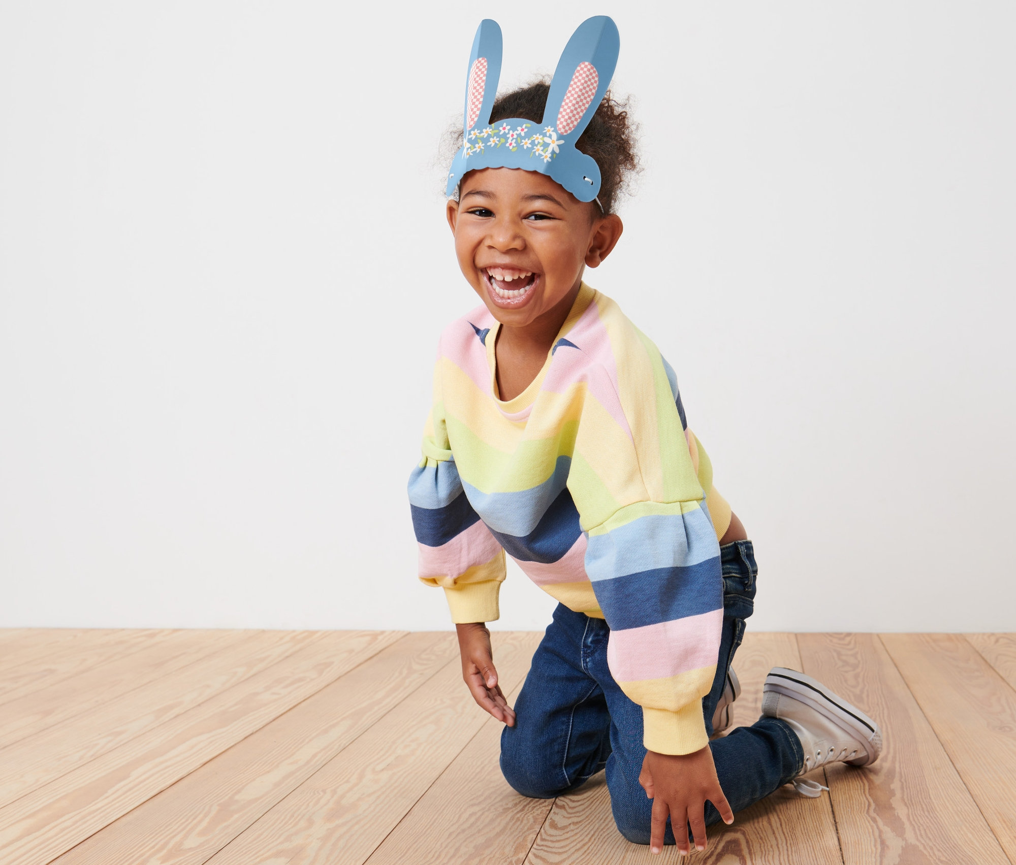 Uśmiechnięte dziecko klęczy na podłodze, ma na sobie papierowe uszy zająca.