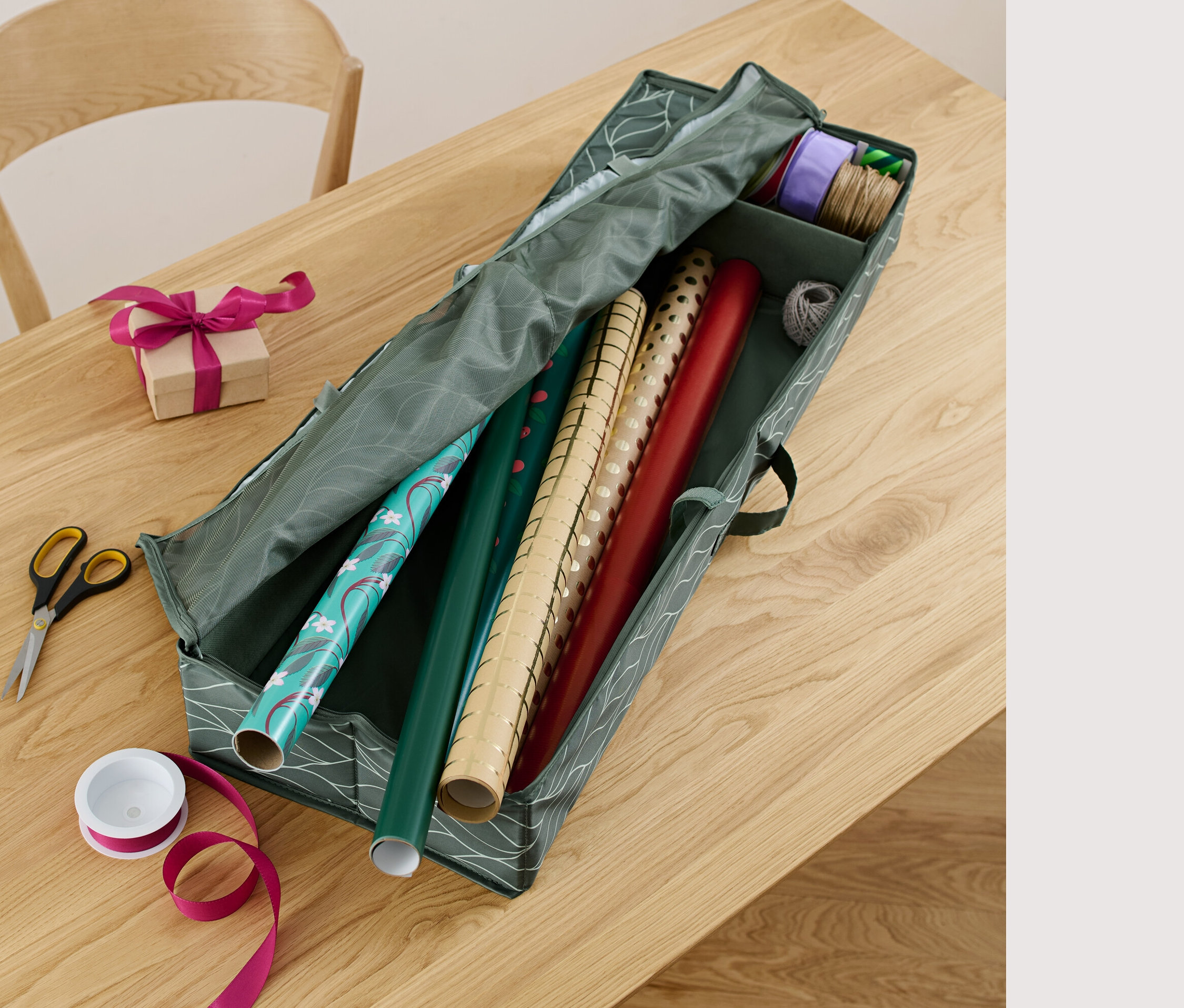 Na stole leżą organizer na papier prezentowy, nożyczki, wstążka i zapakowany prezent.