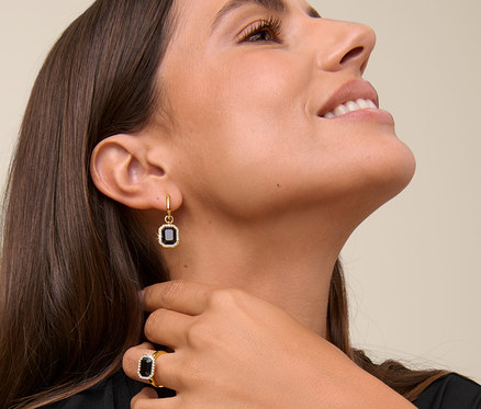 Kobieta uśmiecha się i dotyka szyi, ma na sobie naszyjnik Royal Black, kolczyki koła Royal Black i pierścionek Royal Black.