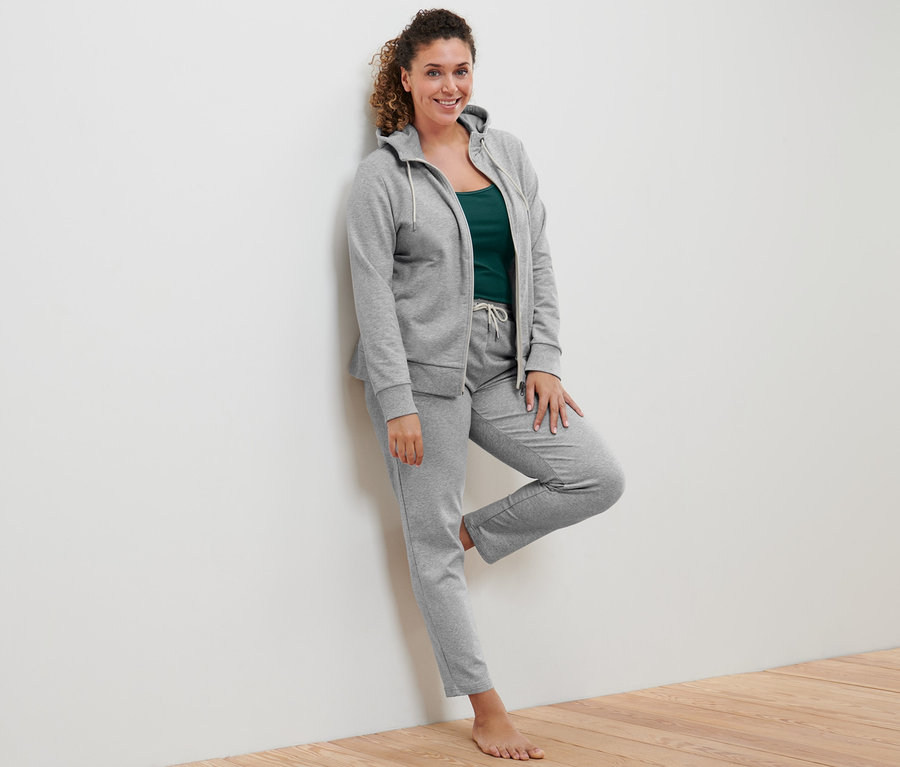 Kobieta oparta o ścianę ma na sobie szarą bluzę z dzianiny dresowej, topy (2 sztuki) w kolorze zielonym i białym oraz szare spodnie dresowe.