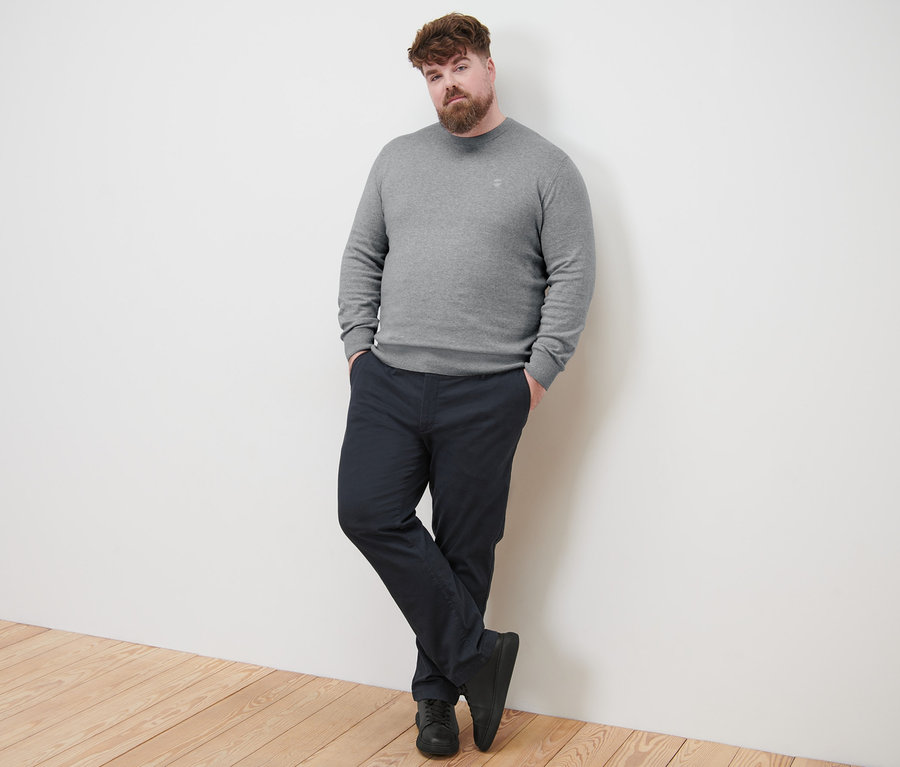 Mężczyzna opiera się o ścianę, ubrany w szary sweter z okrągłym wycięciem i granatowe spodnie chinosy.