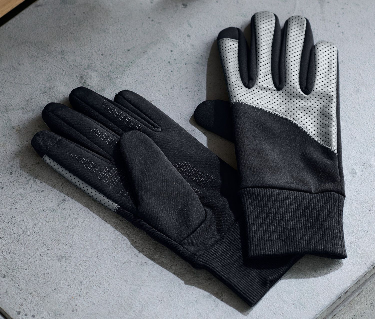 Dwie pary wiatroszczelnych rękawic leżą na szarej powierzchni obok słuchawek i butelki z wodą.