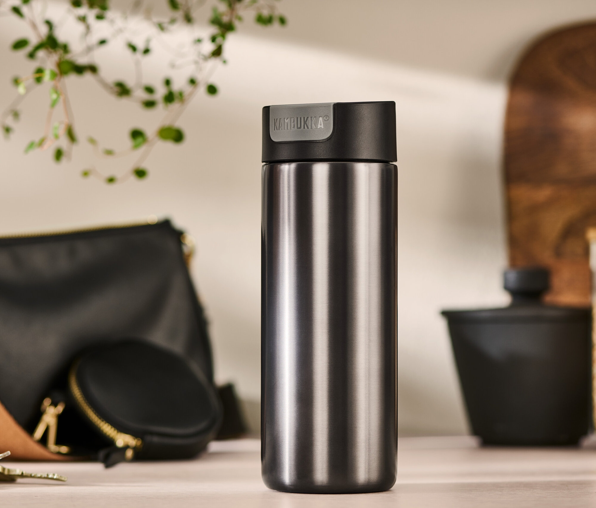 Szary kubek termiczny Kambukka o pojemności 500 ml stoi na stole obok torebki, etui na monety i kluczy.