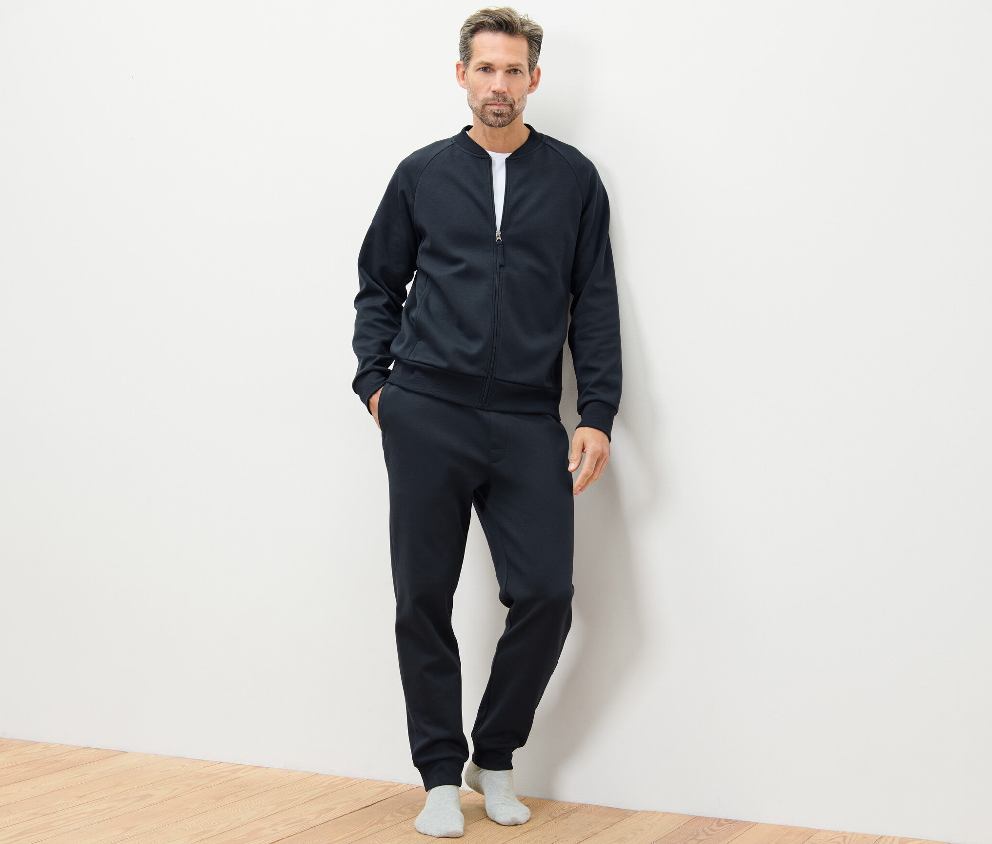 Mężczyzna pozuje na drewnianej podłodze w granatowym dresie domowym.